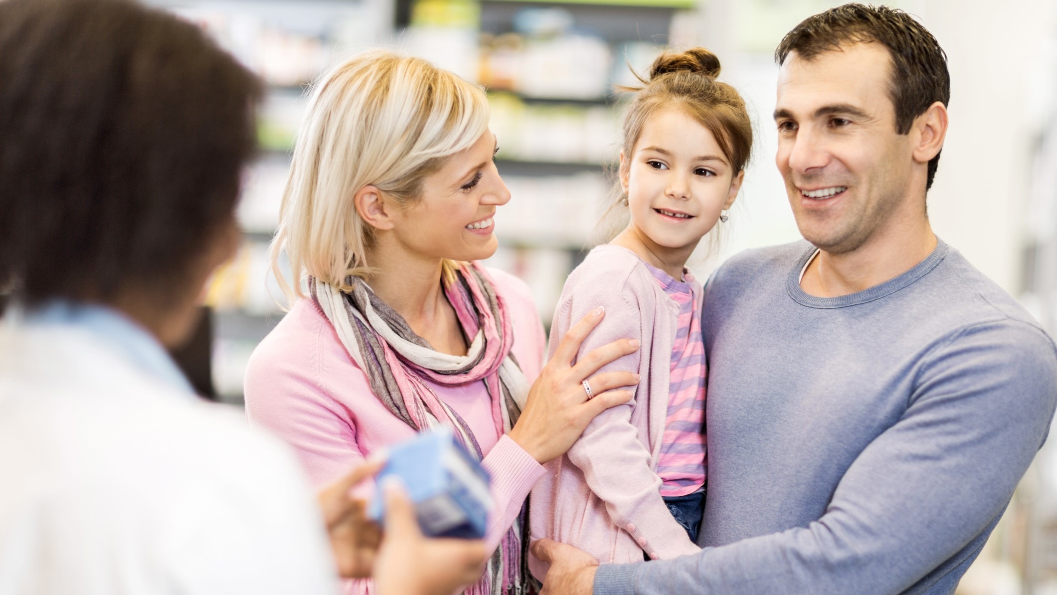 A man and woman have a discussion with a pharmacist.  The man is holding a small child in his arms.  The background is out of focus.